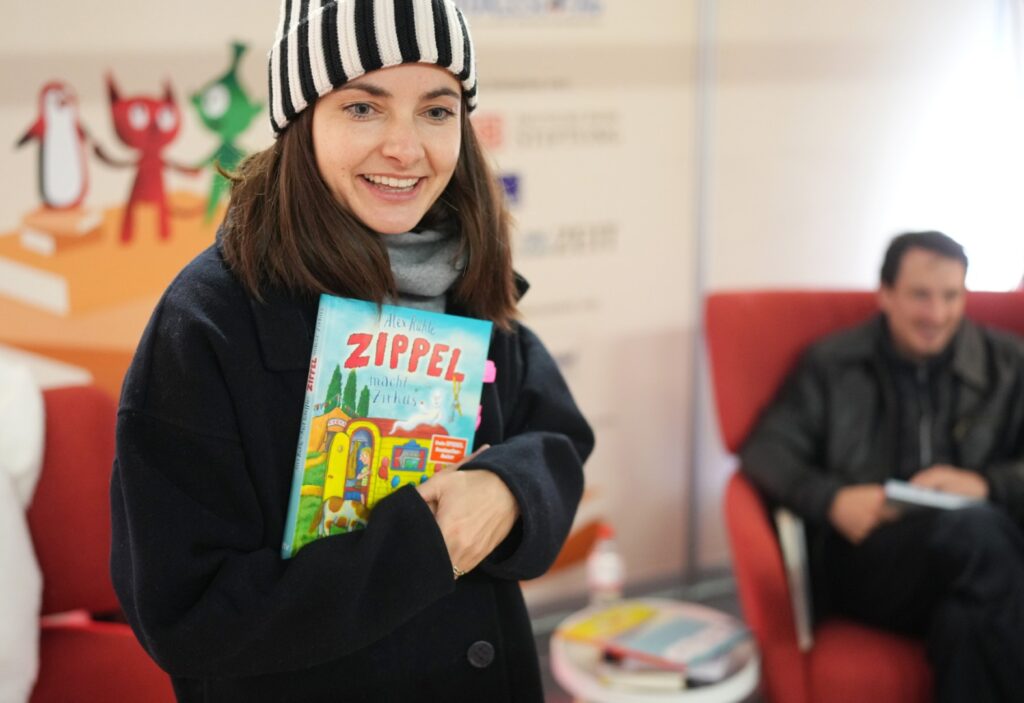 Paula Schramm, Lesebotschafterin und Moderatorin, steht im dicken Wintermantel mit Wollmütze im Vorlesebahnhof der DB. In der Hand hält sie ein Vorlesebuch, das sie gleich vorlesen wird. im Hintergrund ist es fröhlich dekoriert, passend zum Bundesweiten Vorlesetag. Laurence Rupp sitzt hinter ihr auf einem roten Sessel. copyright: Deutsche Bahn Stiftung / Oliver Lang