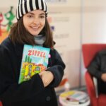 Paula Schramm, Lesebotschafterin und Moderatorin, steht im dicken Wintermantel mit Wollmütze im Vorlesebahnhof der DB. In der Hand hält sie ein Vorlesebuch, das sie gleich vorlesen wird. im Hintergrund ist es fröhlich dekoriert, passend zum Bundesweiten Vorlesetag. Laurence Rupp sitzt hinter ihr auf einem roten Sessel. copyright: Deutsche Bahn Stiftung / Oliver Lang