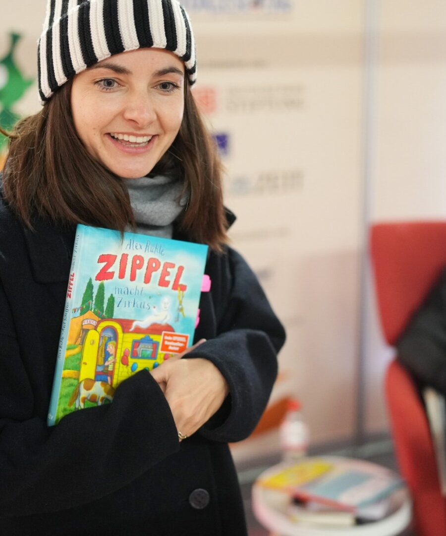 Paula Schramm, Lesebotschafterin und Moderatorin, steht im dicken Wintermantel mit Wollmütze im Vorlesebahnhof der DB. In der Hand hält sie ein Vorlesebuch, das sie gleich vorlesen wird. im Hintergrund ist es fröhlich dekoriert, passend zum Bundesweiten Vorlesetag. Laurence Rupp sitzt hinter ihr auf einem roten Sessel. copyright: Deutsche Bahn Stiftung / Oliver Lang