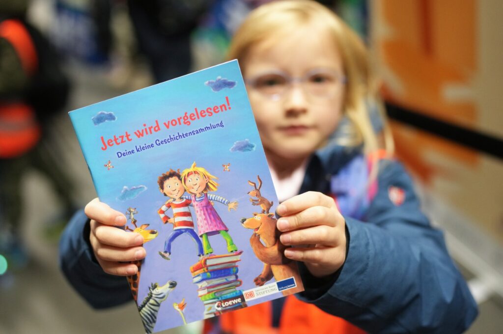 ein Mädchen hält ein Buchheft "Jetzt wird vorgelesen!" der Deutsche Bahn Stiftung in der Hand