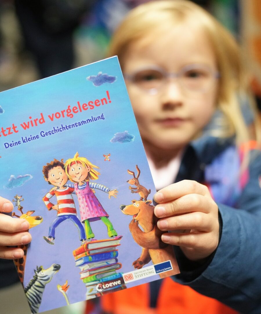 ein Mädchen hält ein Buchheft "Jetzt wird vorgelesen!" der Deutsche Bahn Stiftung in der Hand