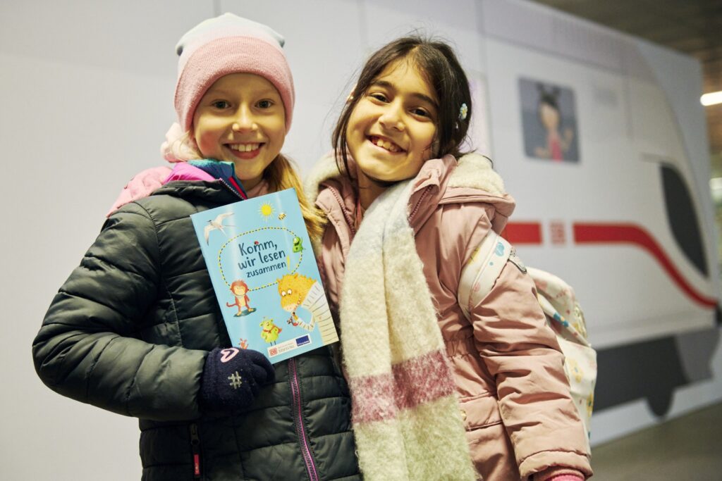 Zwei Mädchen stehend lachend vor einem ICE und halten ein Buch in der Hand