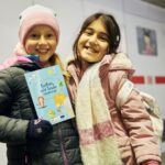 Zwei Mädchen stehend lachend vor einem ICE und halten ein Buch in der Hand