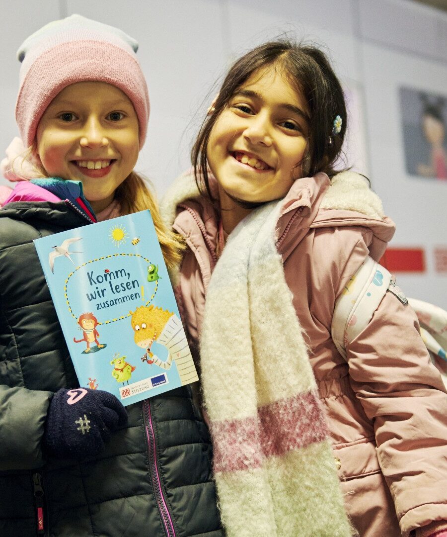 Zwei Mädchen stehend lachend vor einem ICE und halten ein Buch in der Hand
